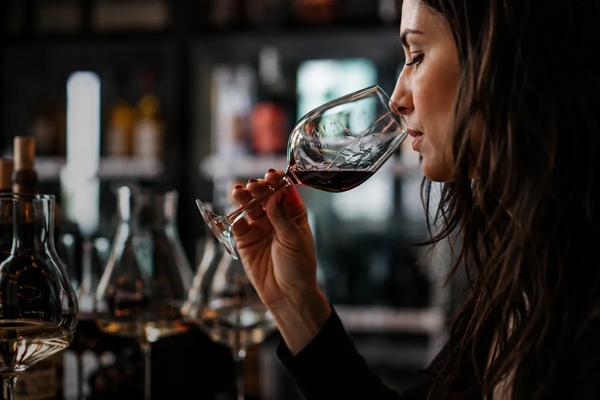 Mulher de perfil levando uma taça de vinho tinto ao nariz, em um balcão de bar