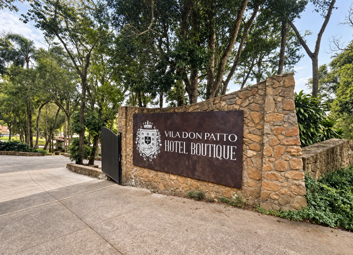Muro de pedra com a placa VILA DON PATTO HOTEL BOUTIQUE em letras douradas, ao lado de um portão, com árvores altas atrás.