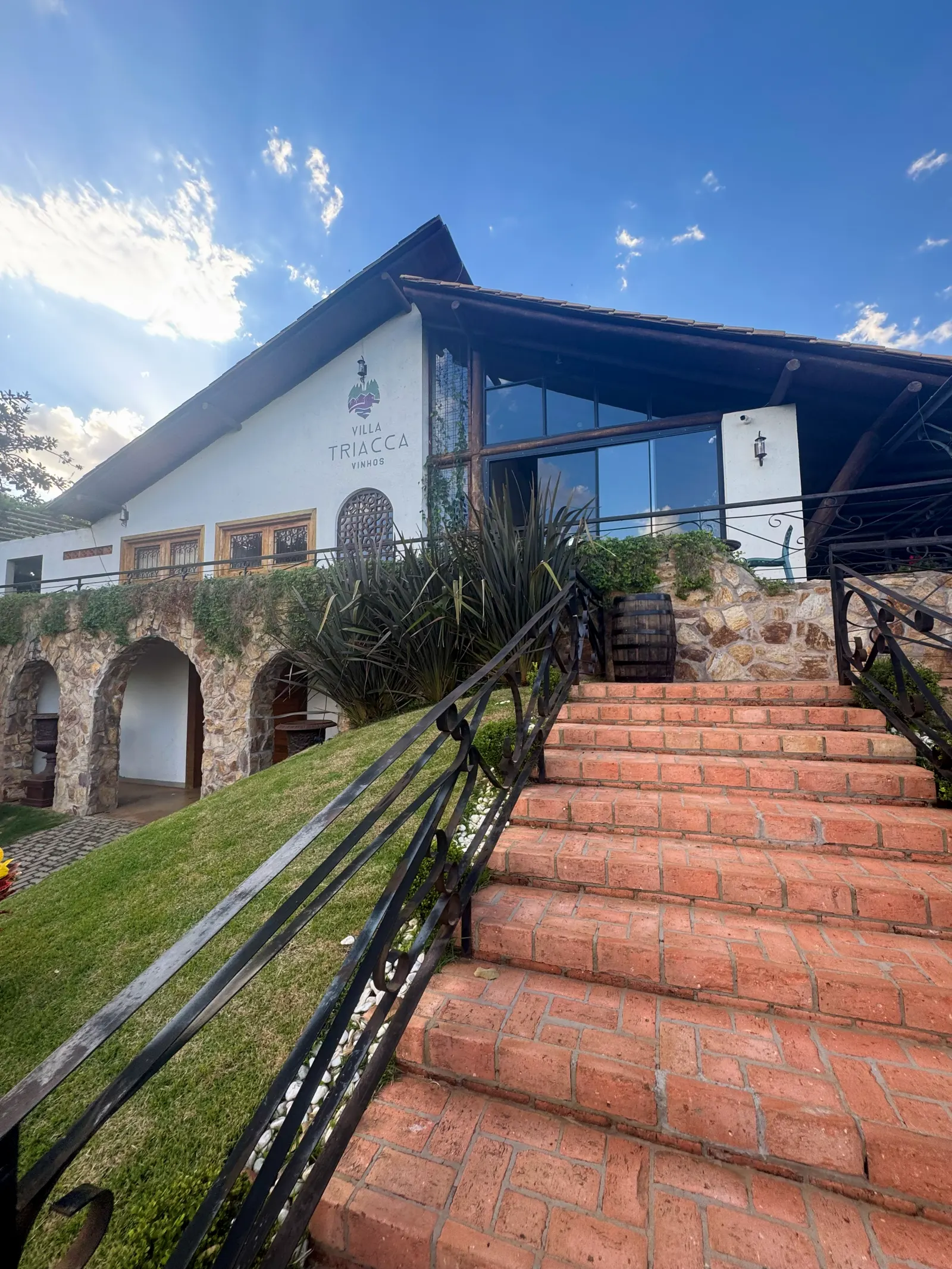 Fachada de pedra branca e vidro da Villa Triacca, com uma escada larga de tijolo vermelho subindo até a entrada e arcos no térreo.