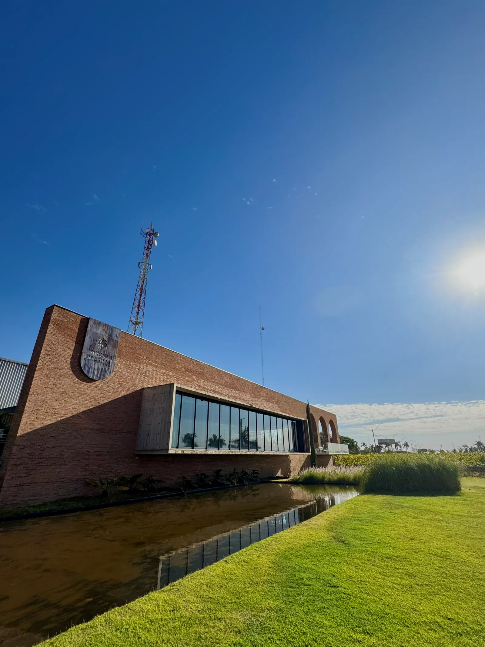 Edifício moderno de tijolo aparente, com uma faixa horizontal de vidro e cobertura inclinada, refletido num espelho d'água. Céu azul limpo e gramado à frente.